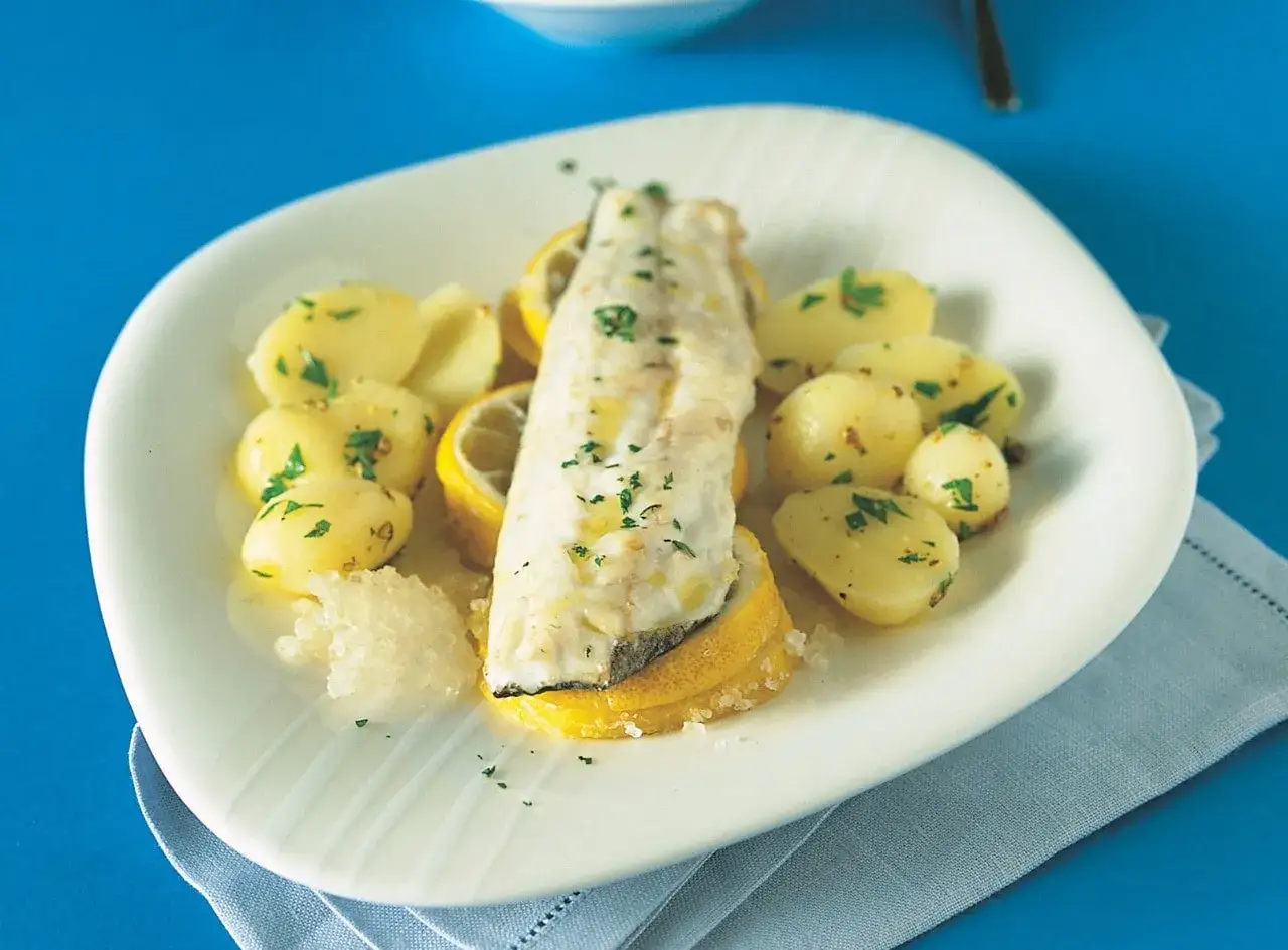 Filetti di branzino in padella, cotti alla perfezione, serviti con patate novelle e spicchi di limone su un piatto bianco.