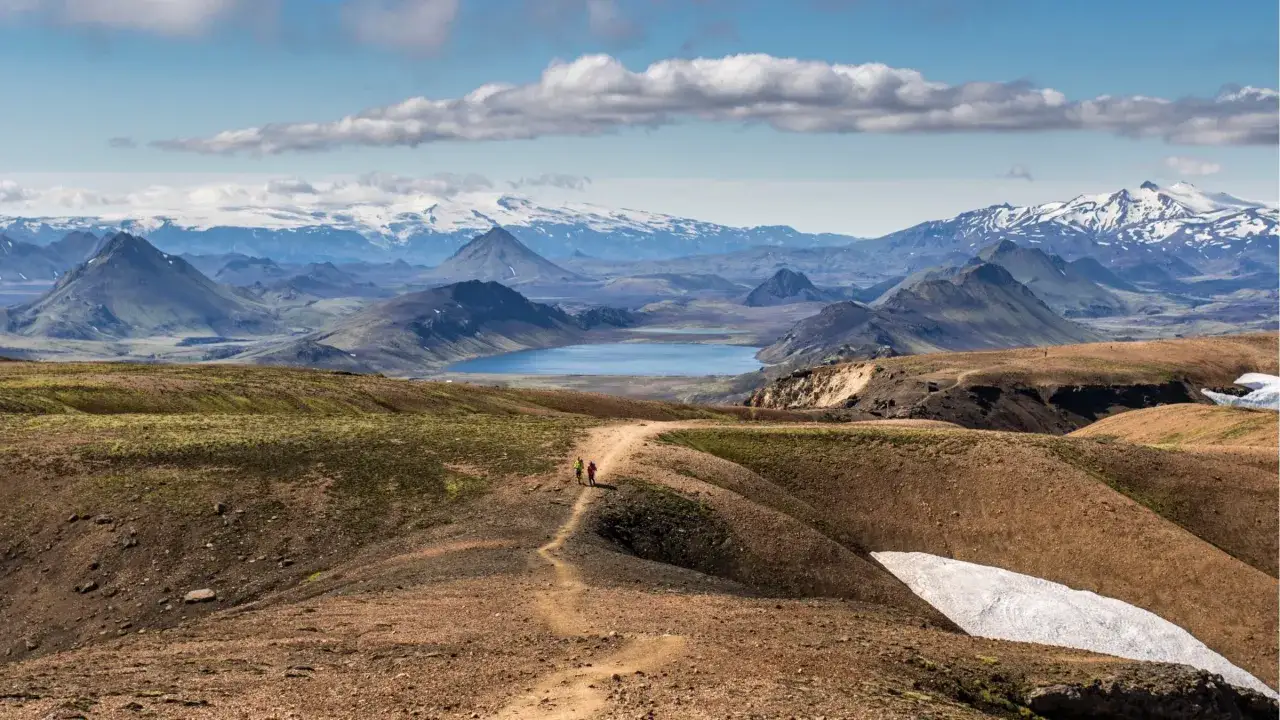 Zwei Wanderer auf einem Pfad in einer bergigen Landschaft mit einem See und schneebedeckten Gipfeln im Hintergrund. Perfekt f&uuml;r mehrtageswanderungen Europa.