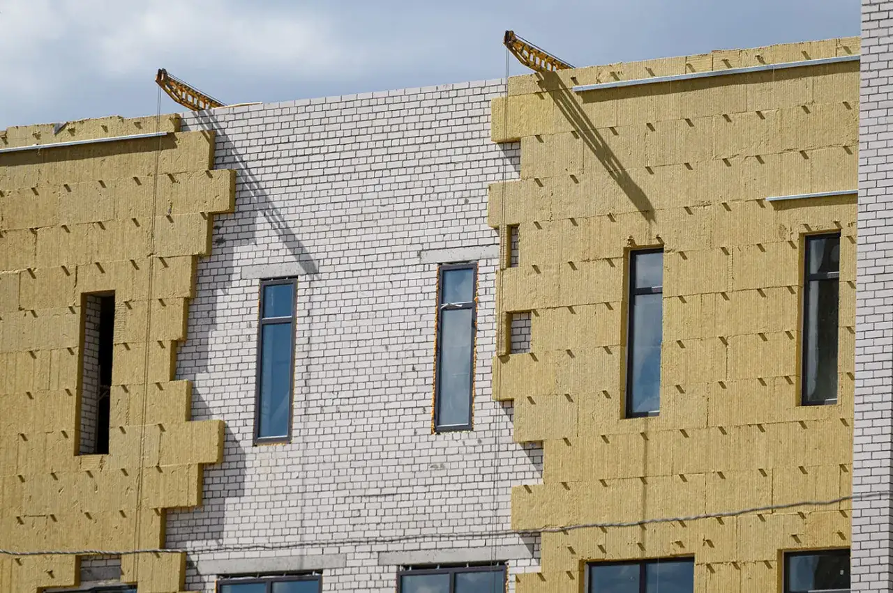 Fachadas en construcci&oacute;n con aislamiento amarillo y ladrillo blanco. Se aprecian ventanas y gr&uacute;as en la parte superior.