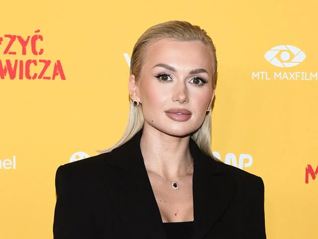 Kobieta z blond włosami i makijażem, w czarnej marynarce, na tle z napisem 