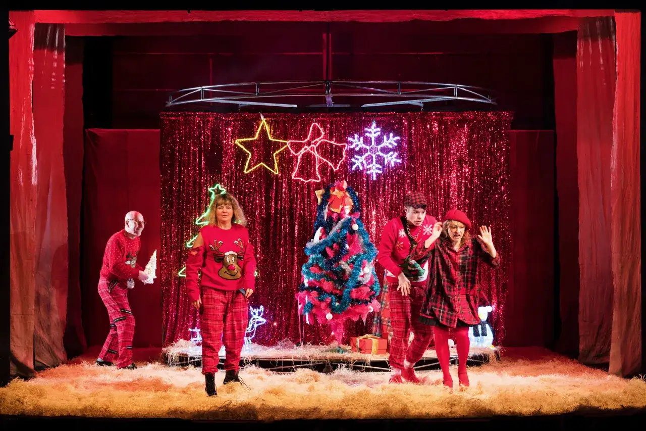 Wesoły spektakl teatralny dla dzieci z motywem świątecznym. Aktorzy w czerwonych strojach, choinka i błyszczące tło tworzą magiczną atmosferę.