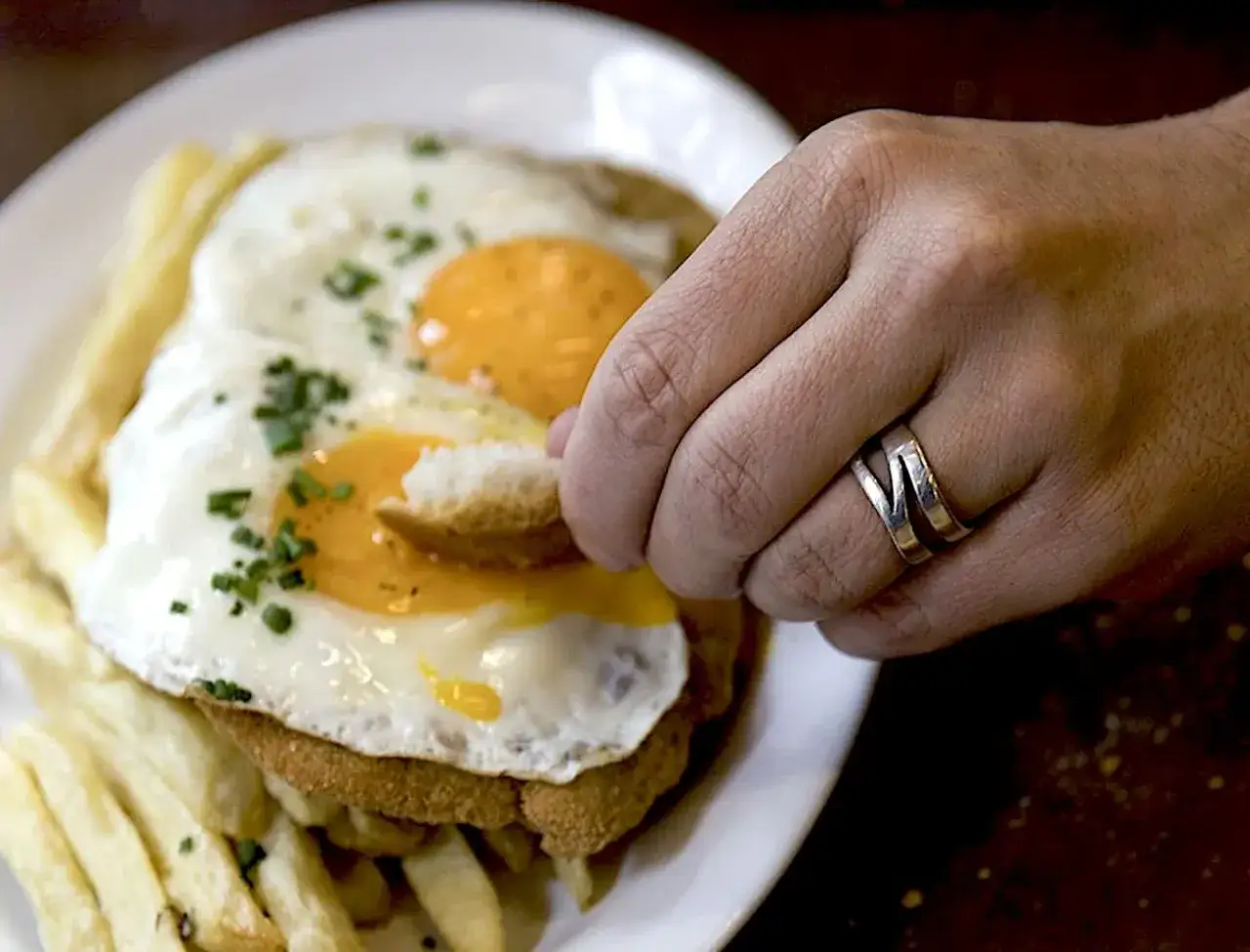 Un trozo de pan se hunde en un huevo frito sobre una milanesa a caballo, acompa&ntilde;ada de papas fritas.