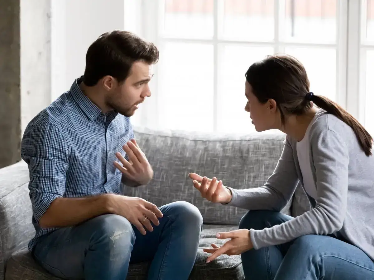 Pareja discutiendo en un sof&aacute;. &Eacute;l gesticula con las manos, ella lo mira fijamente. Una escena de **amor posesivo** que se desmorona.