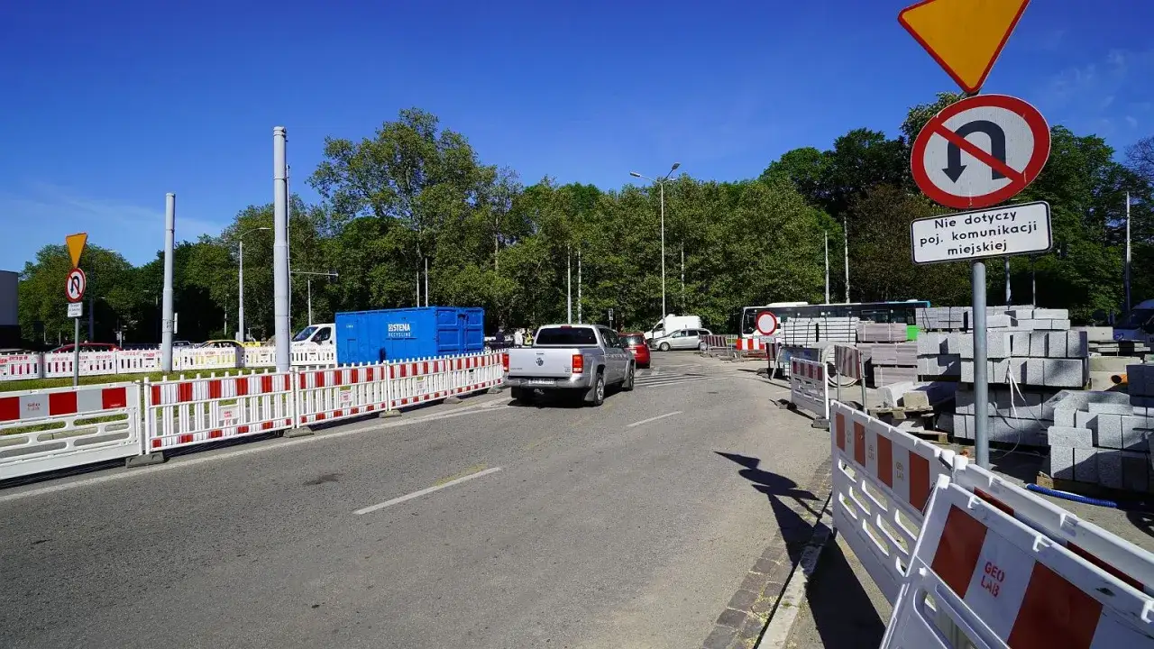 Srebrny pickup wykonuje zawracanie na skrzyżowaniu, omijając tymczasowe bariery budowlane i stosy kostki brukowej.
