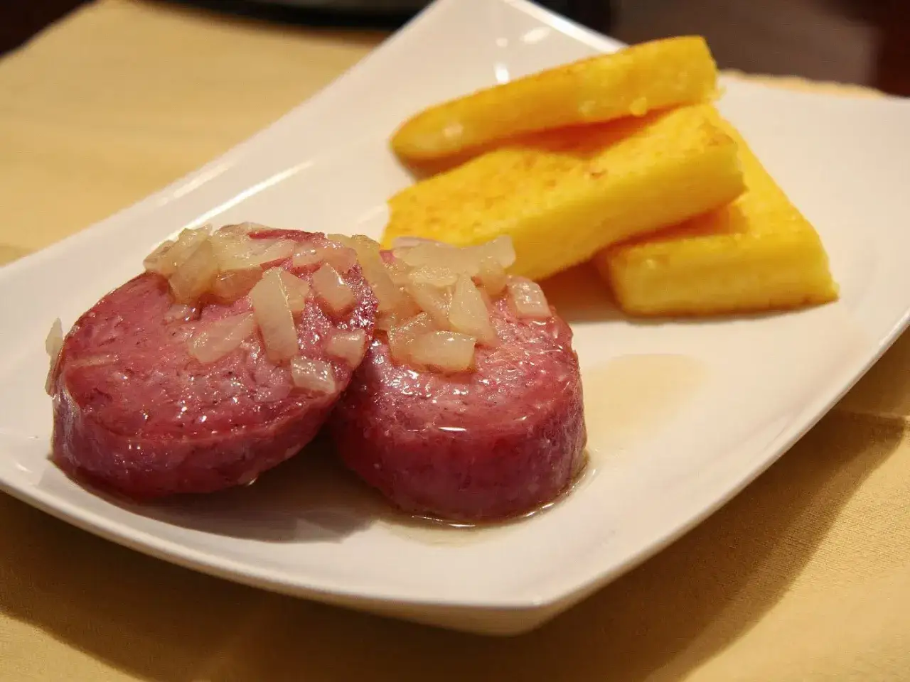 Fette di salame in gravidanza, condite con cipolla, servite con bastoncini di polenta dorata su un piatto bianco.