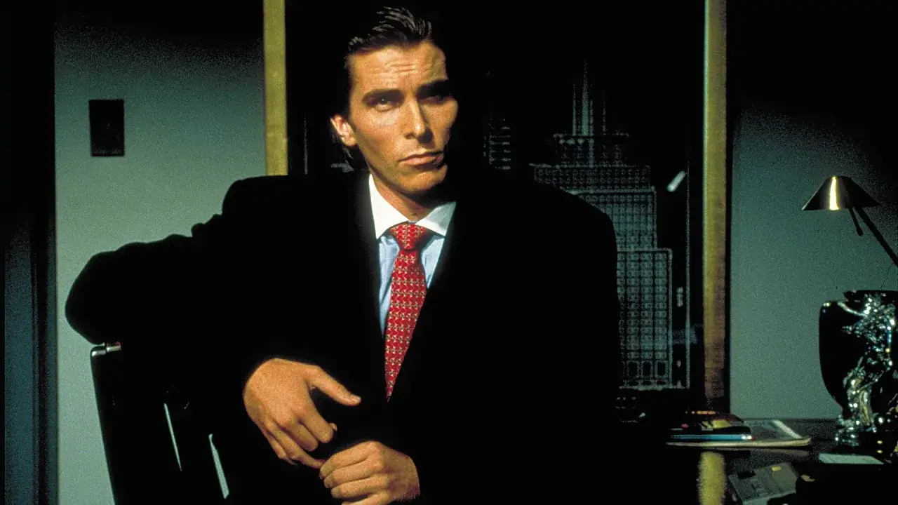 Patrick Bateman, från American Psycho, sitter i en mörk miljö, klädd i kostym och slips.