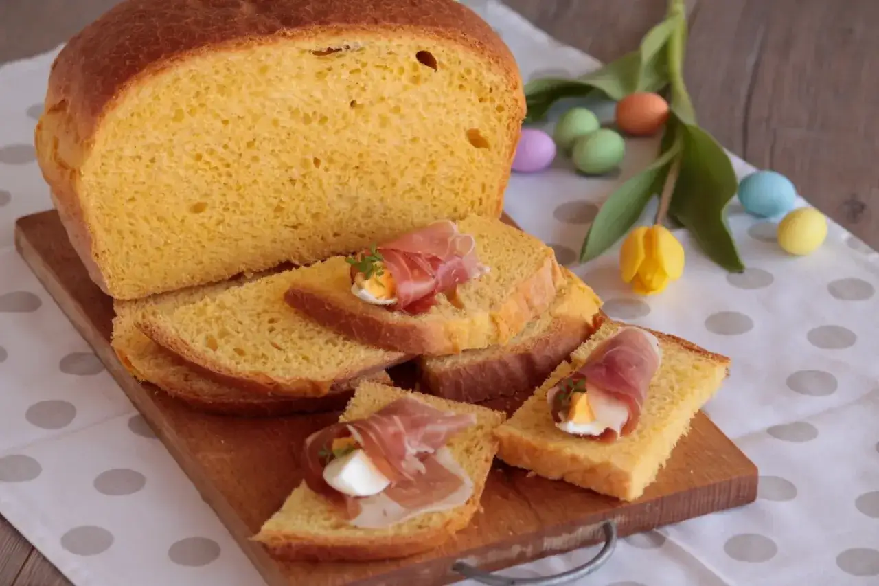 Fetta di torta al formaggio umbra ricetta originale, guarnita con uovo sodo e prosciutto crudo. Uova colorate e tulipano sullo sfondo.