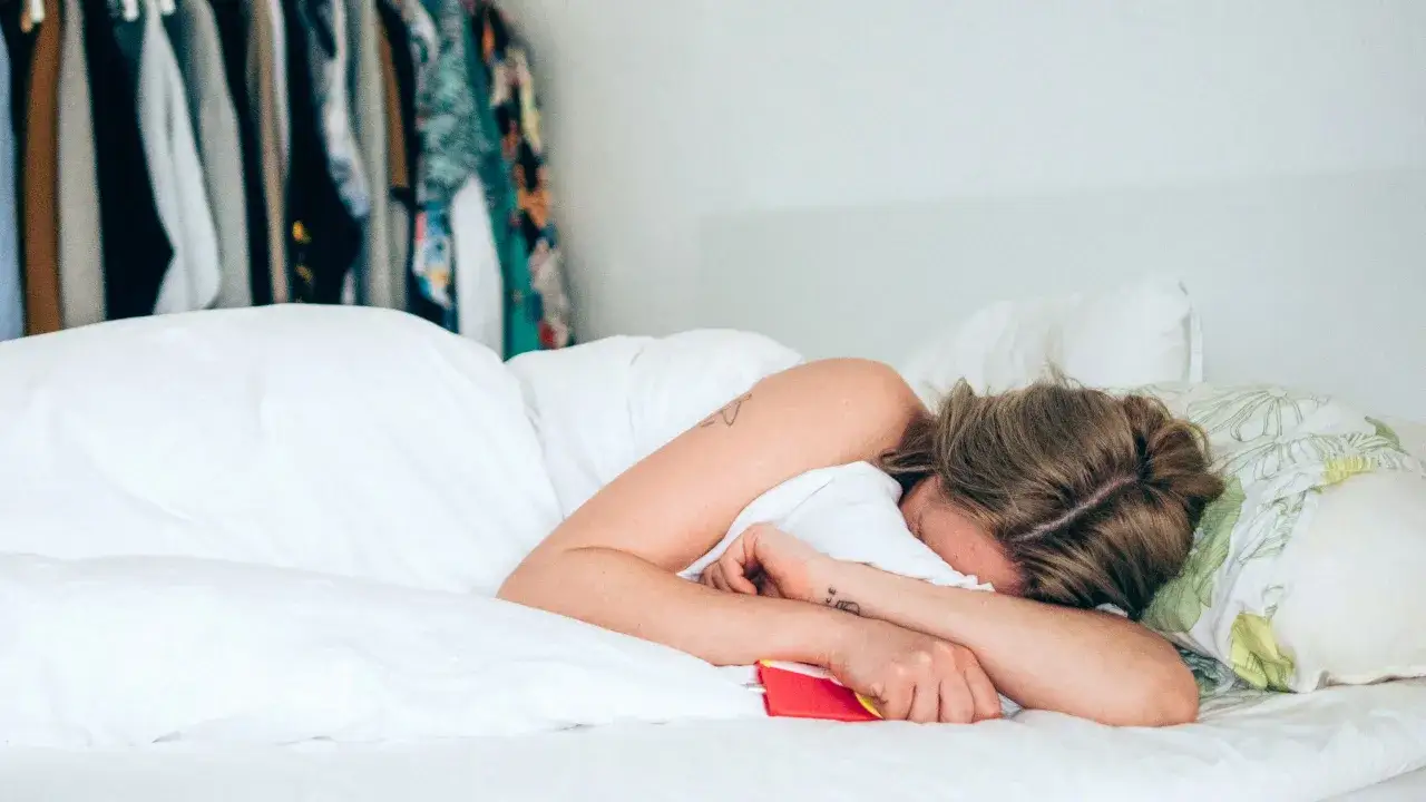 Frau liegt im Bett, Arme um den Kopf geschlungen, ein rotes Handy liegt daneben. Sie leidet unter Übelkeit vor Periode.