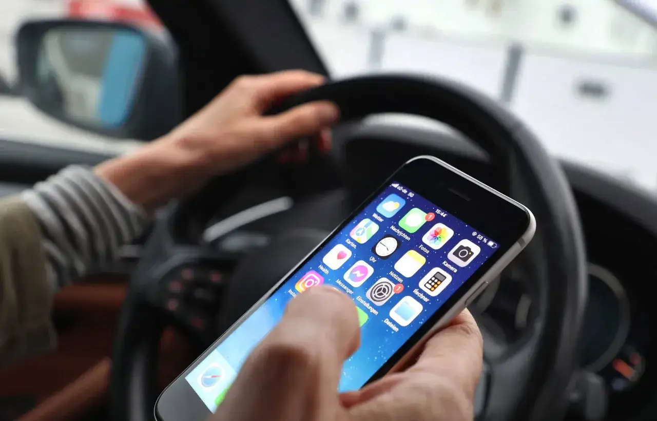 Handy am Steuer: Ein Finger tippt auf dem Smartphone, w&auml;hrend die H&auml;nde das Lenkrad halten. Das kann eine teure Strafe bedeuten.
