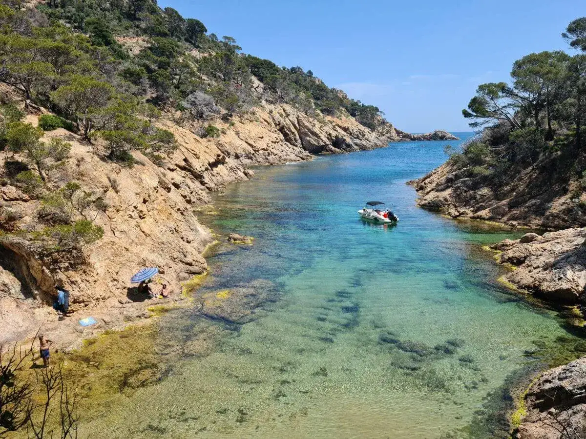 Aguas cristalinas en las calas de Tossa de Mar. Una pequeña barca descansa en la bahía, mientras unas personas disfrutan del sol en la orilla rocosa.