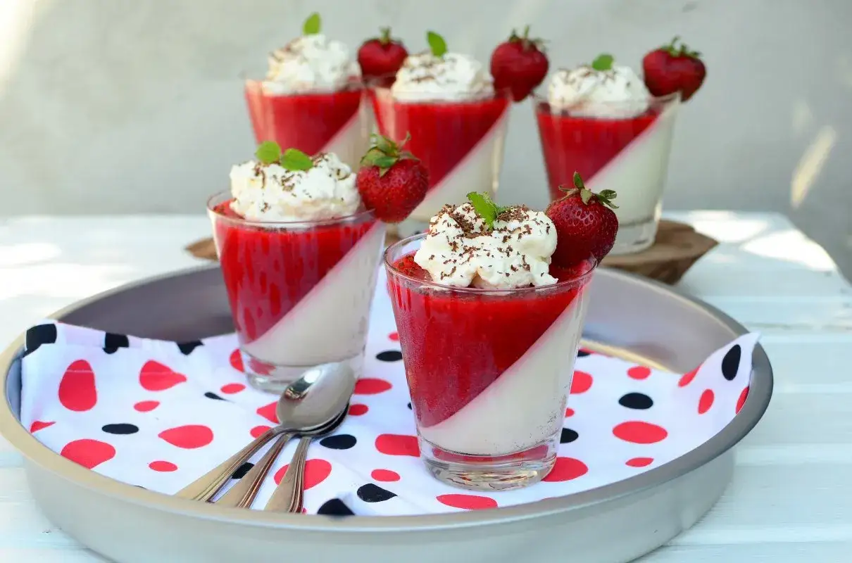Panna cotta z truskawkami, udekorowana bitą śmietaną, świeżymi owocami i listkiem mięty. Letni deser w szklanych pucharkach.