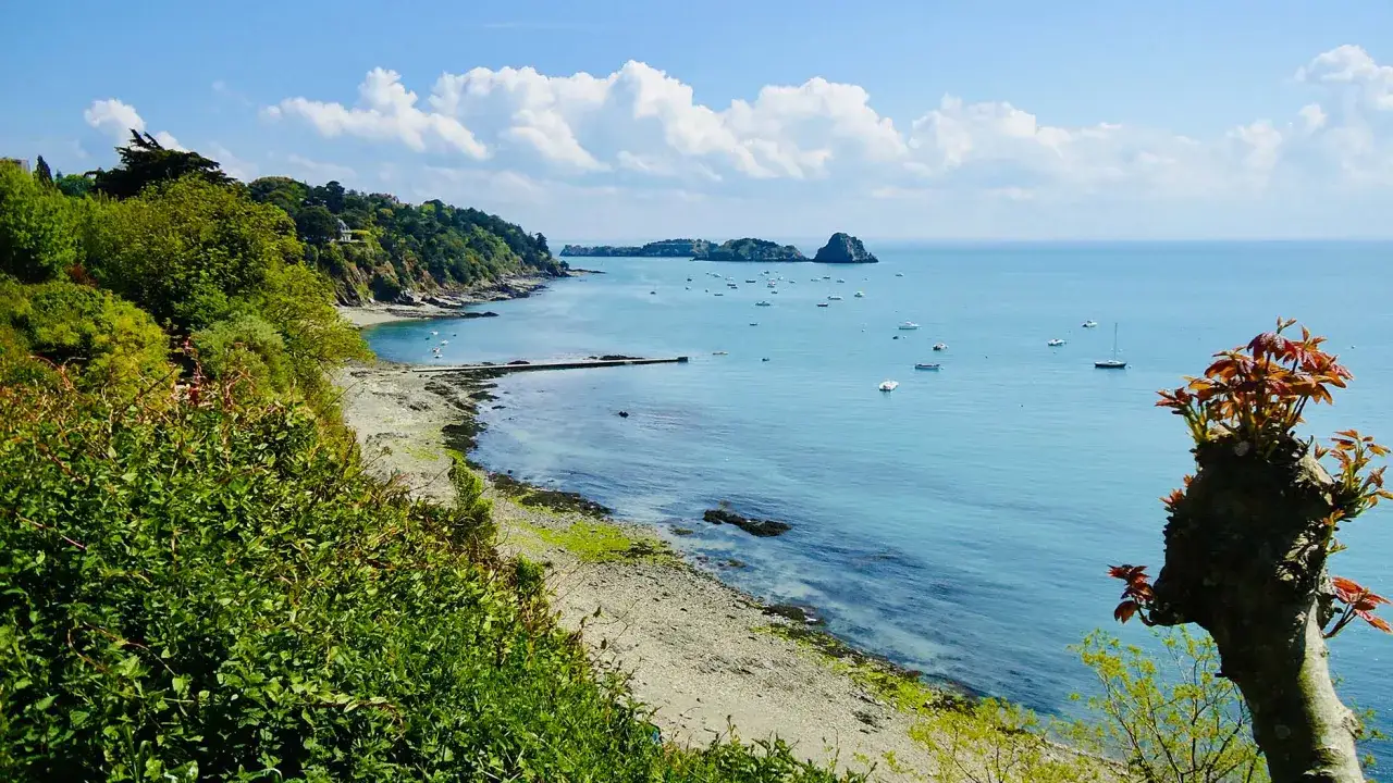 Vue sur la côte verdoyante du pays malouin, avec des bateaux sur la mer bleue et un ciel d'été nuageux.
