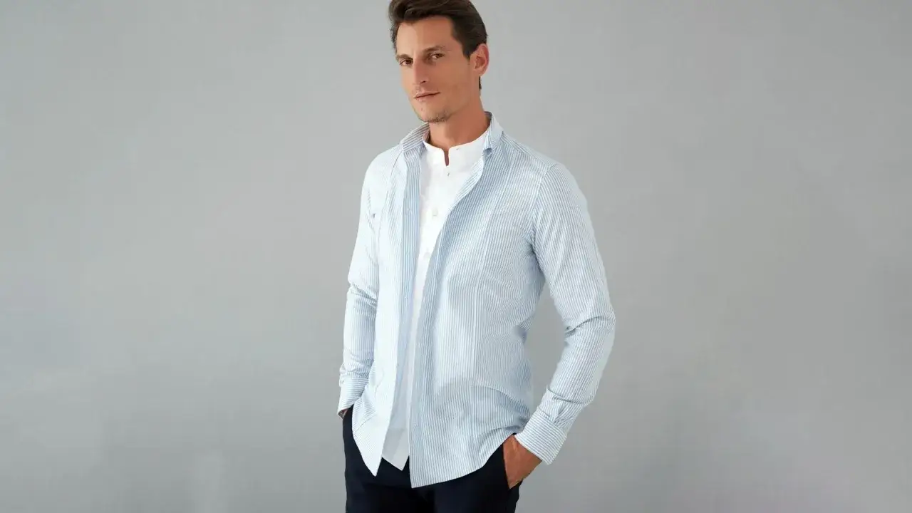 Uomo con outfit camicia a righe azzurre e bianche, indossata aperta su una t-shirt bianca. Mani nelle tasche.