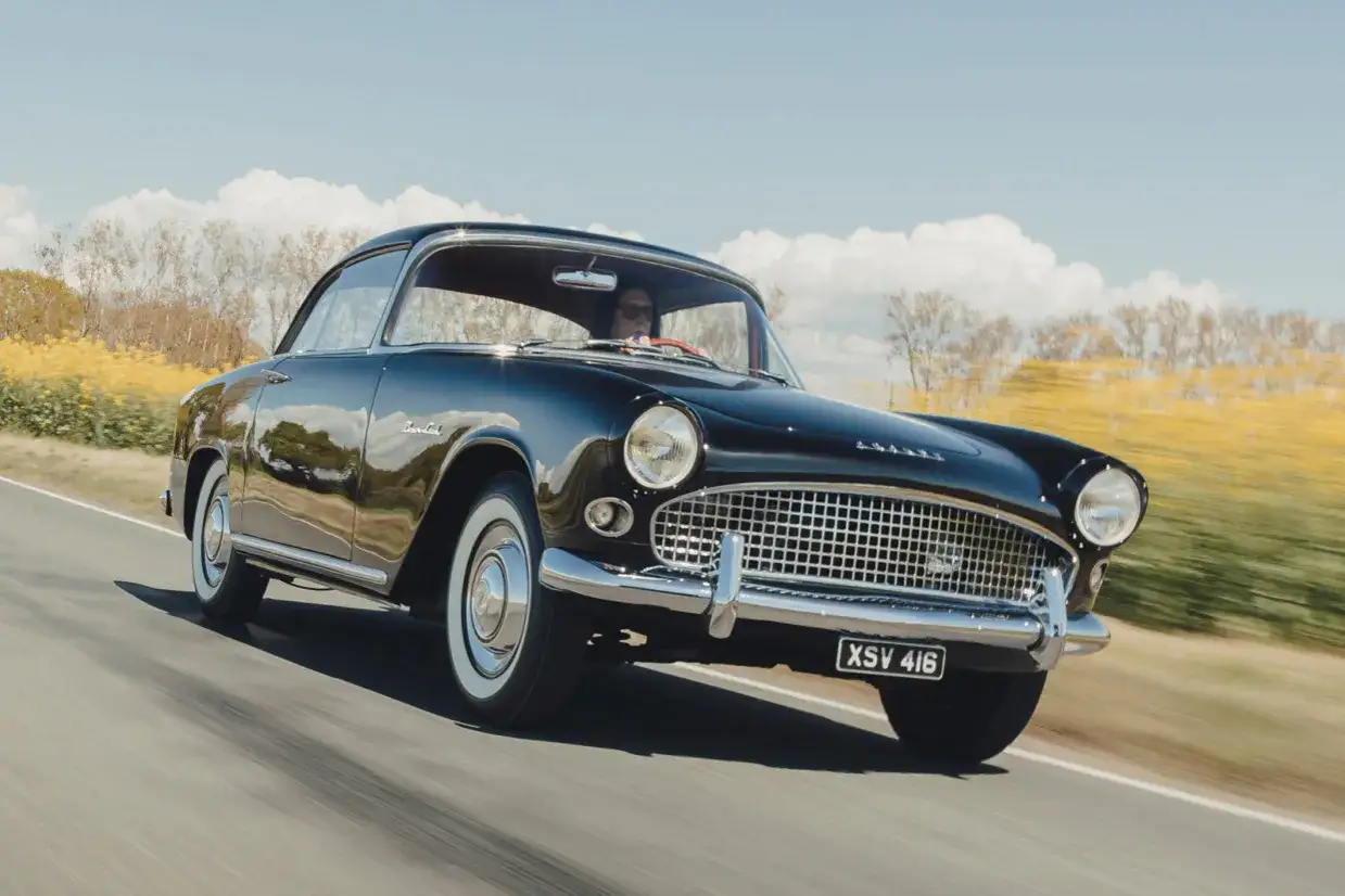 Svart, blank simca bil k&ouml;r l&auml;ngs en landsv&auml;g med gula f&auml;lt och tr&auml;d i bakgrunden.