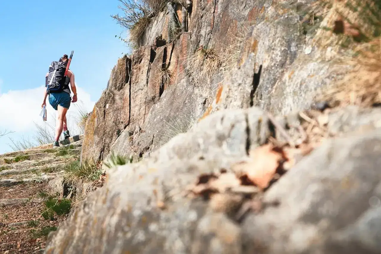 Une randonneuse avec un grand sac &agrave; dos monte un sentier montagneux rocailleux sous un ciel bleu clair.