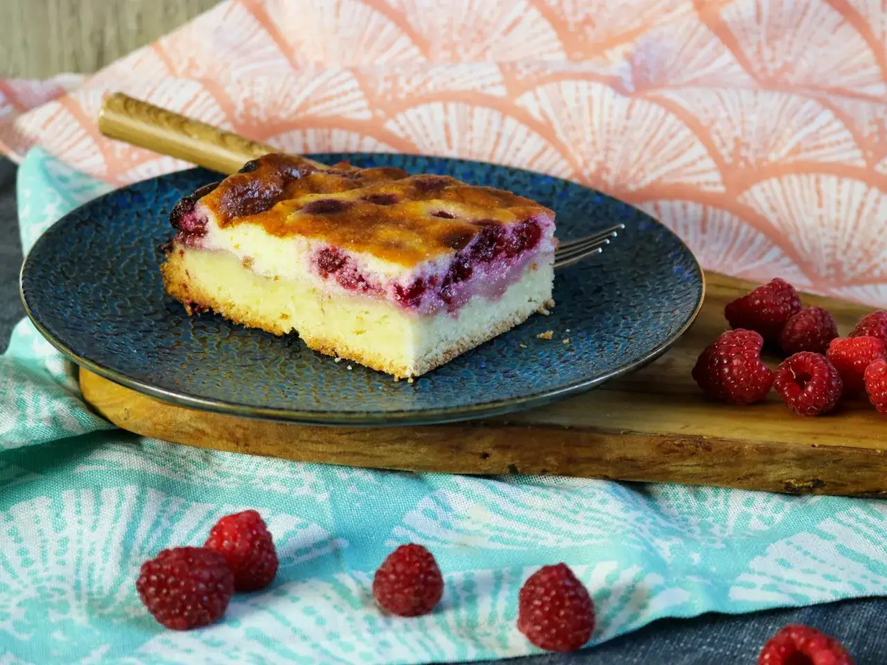 Ein St&uuml;ck Himbeer Schmand Kuchen auf einem blauen Teller, daneben frische Himbeeren.