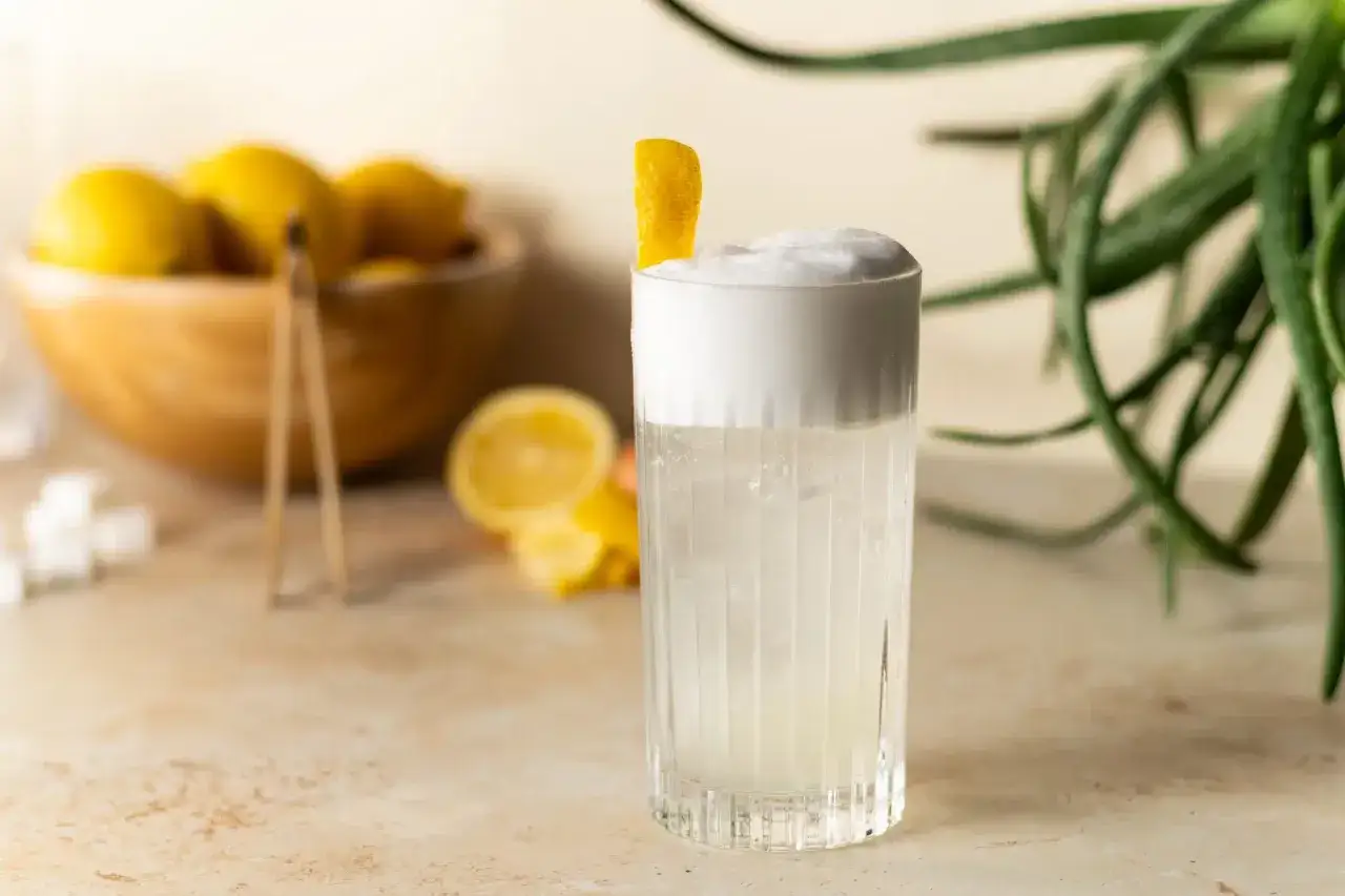 Un verre de gin fizz mousseux, garni d'un zeste de citron, avec des citrons et des morceaux de sucre en arrière-plan. Parfait pour une recette de gin fizz rafraîchissante.