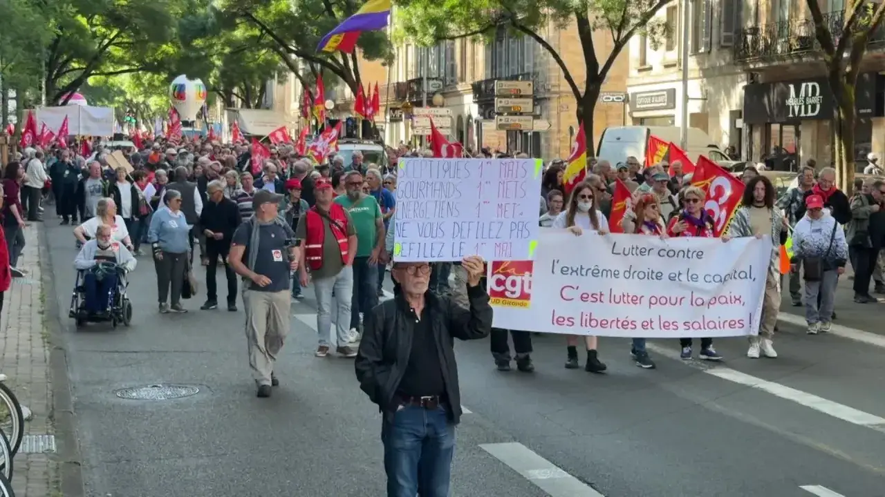 Manifestation du 1er mai avec drapeaux rouges et banderoles. Une foule marche dans la rue, certains portent des pancartes.
