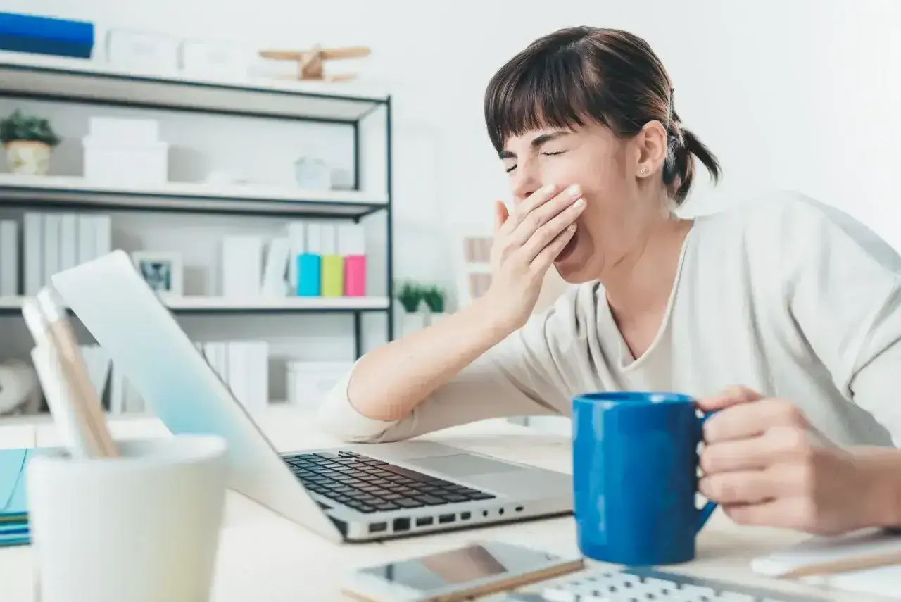 Kobieta ziewająca przy laptopie, z kubkiem kawy. Może to być jeden z objawów psychicznych niewydolności nerek, jak zmęczenie i apatia.