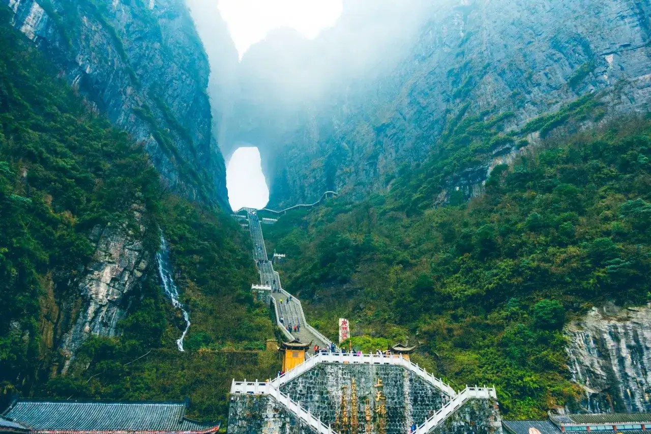 Escaleras ascienden hacia la **puerta del cielo china**, un arco natural en la montaña cubierto de niebla.