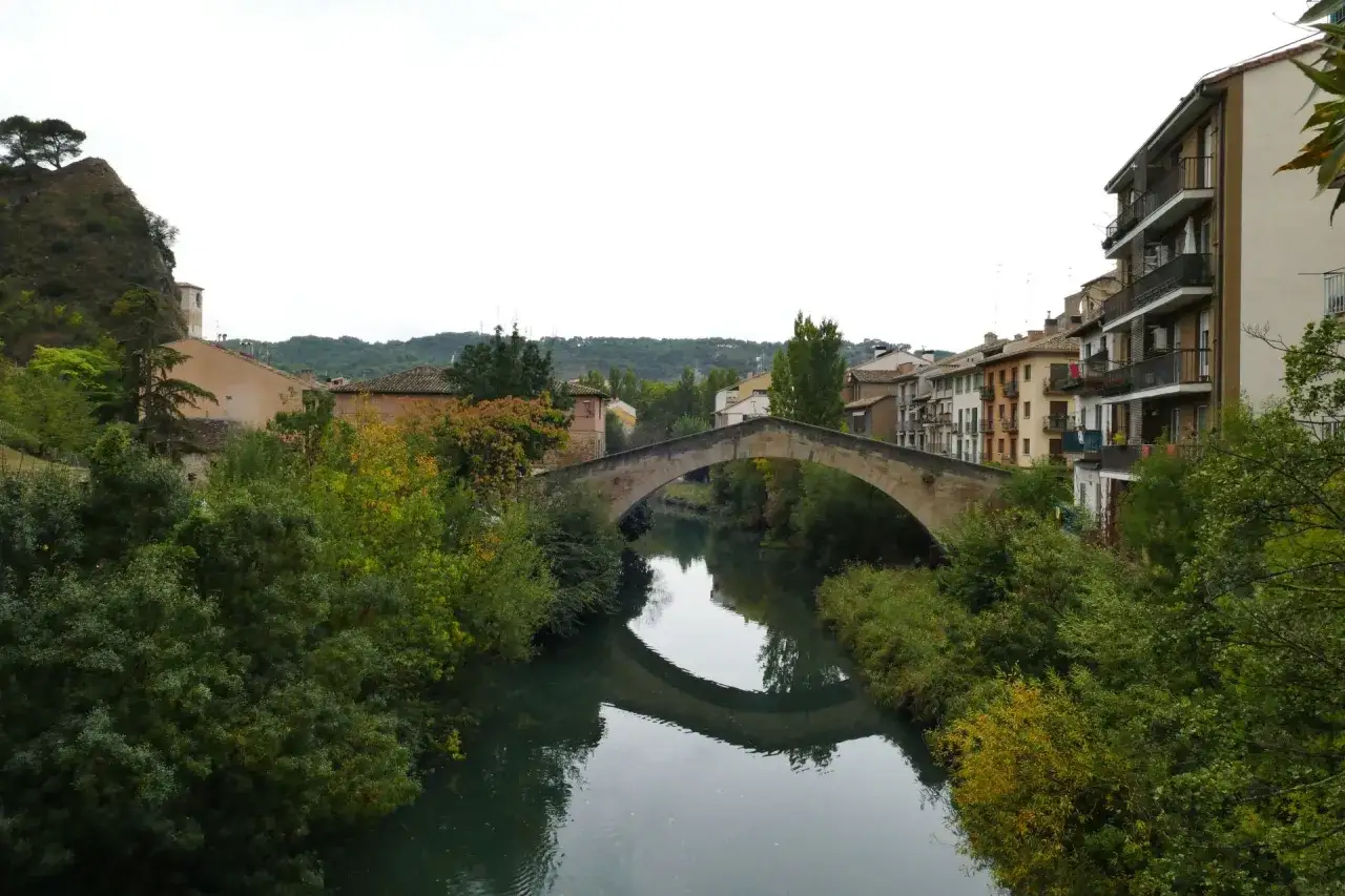 El puente de la cárcel de Estella cruza un río sereno, reflejando su arco de piedra entre frondosa vegetación y edificios históricos.