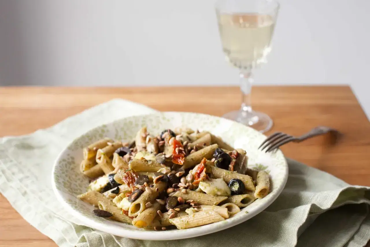Pyszny makaron z pesto i suszonymi pomidorami, ozdobiony pestkami słonecznika i oliwkami, podany na talerzu z zielonym wzorem. Obok kieliszek białego wina.
