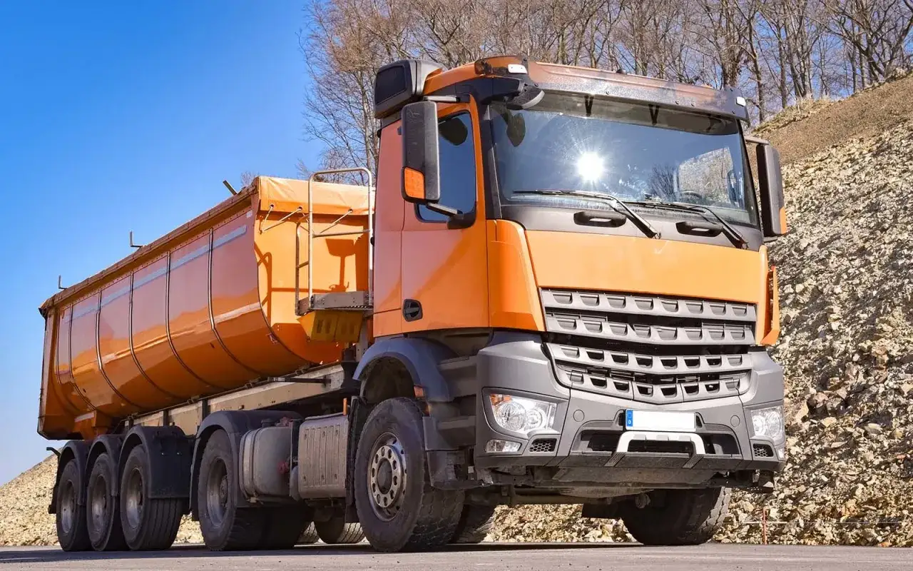 Un camion benne orange, pr&ecirc;t pour le transport, avec un ciel bleu et un tas de gravats en arri&egrave;re-plan. La consommation de ce v&eacute;hicule est importante.