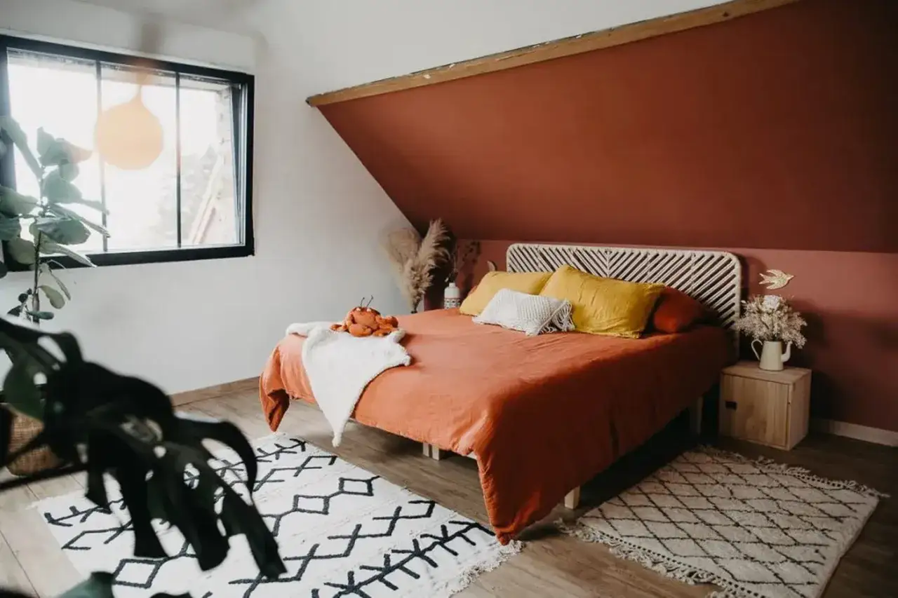 Une chambre couleur terracotta chaleureuse avec un lit douillet, des coussins jaune moutarde et une plante luxuriante.