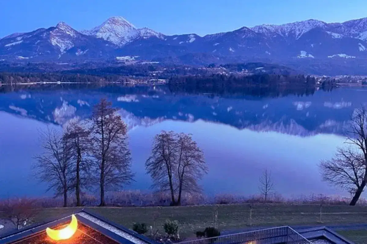 Spokojne jezioro odbija ośnieżone góry w Karyntii, Austria. Na pierwszym planie widać dach z księżycową lampą.