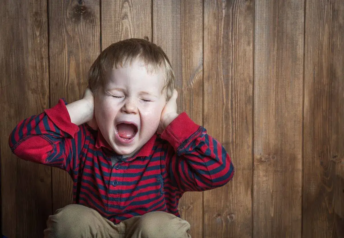 Petit garçon de 5 ans, yeux fermés, criant fort et se bouchant les oreilles, en pleine crise de colère incontrôlable.