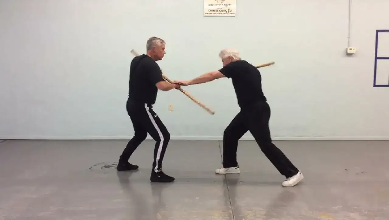 Tv&aring; m&auml;n tr&auml;nar kampsport med pinnar. De st&aring;r mitt emot varandra och h&aring;ller varsin pinne i h&auml;nderna, redo att attackera eller f&ouml;rsvara sig.