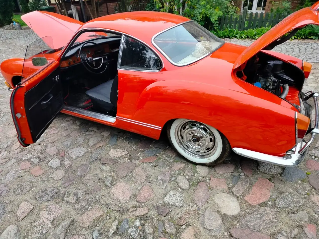 Pomarańczowy Karmann Ghia z otwartymi drzwiami i maską silnika, stojący na brukowanej drodze.