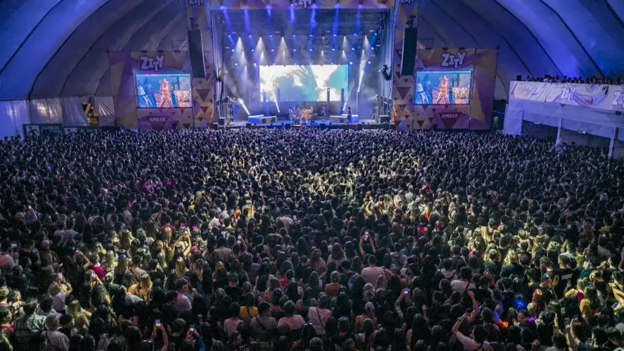 Gran concierto con un aforo masivo en un espacio Zity. Miles de personas disfrutan de la música bajo un techo abovedado, con luces y pantallas gigantes iluminando el escenario.