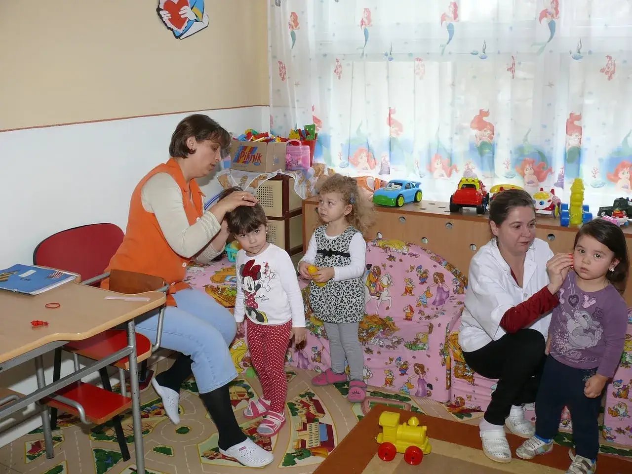 W żłobku, który jest placówką oświatową, opiekunki zajmują się dziećmi. Jedna czesze dziewczynkę, druga bawi się z inną.
