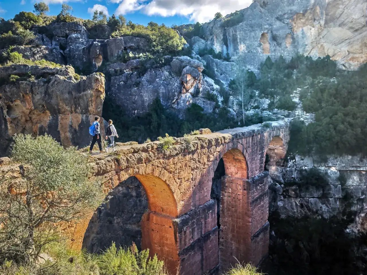 Dos excursionistas cruzan un antiguo acueducto de piedra en Chelva, un pueblo azul rodeado de imponentes formaciones rocosas y vegetación.