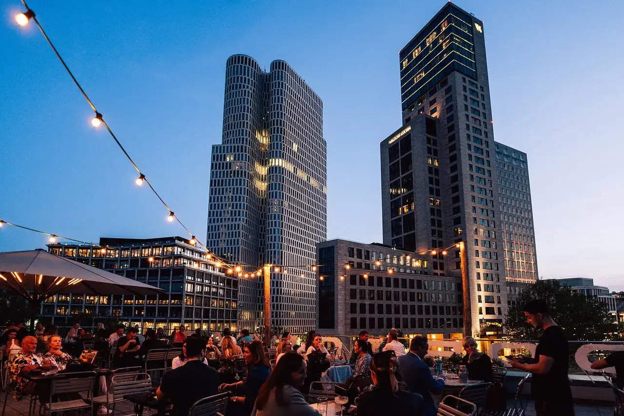 Abendstimmung auf einer Dachterrasse in Berlin. Menschen genießen den Ausblick auf die Skyline, darunter das markante Bikini-Haus. Lichterketten beleuchten die Szene.