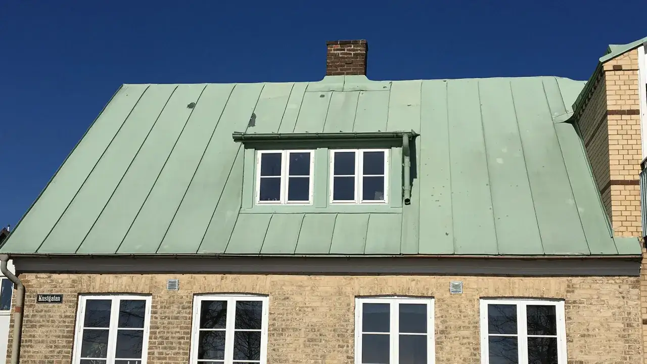 Ett hus med ett gr&ouml;nt pl&aring;ttak gammal stil, tegelv&auml;gg och vita f&ouml;nster under en klarbl&aring; himmel.