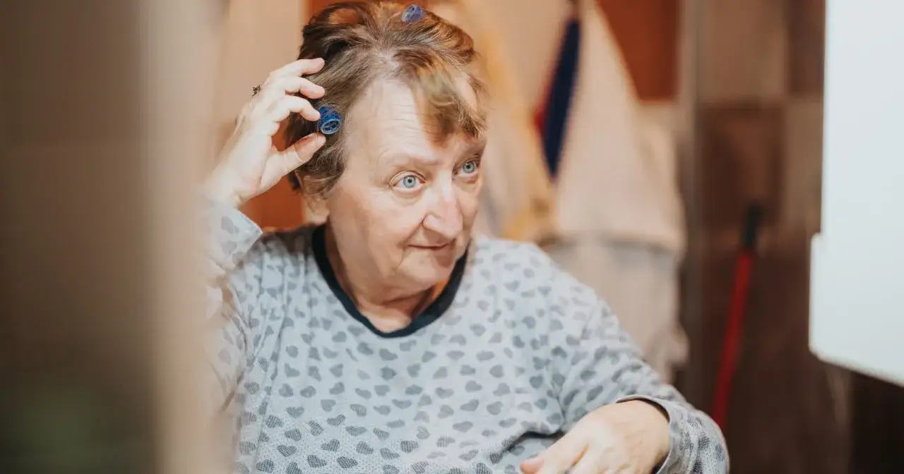 Starsza kobieta z niebieskimi wałkami we włosach, jej spojrzenie zagubione, jakby walczyła z demencją, próbując ułożyć fryzurę.