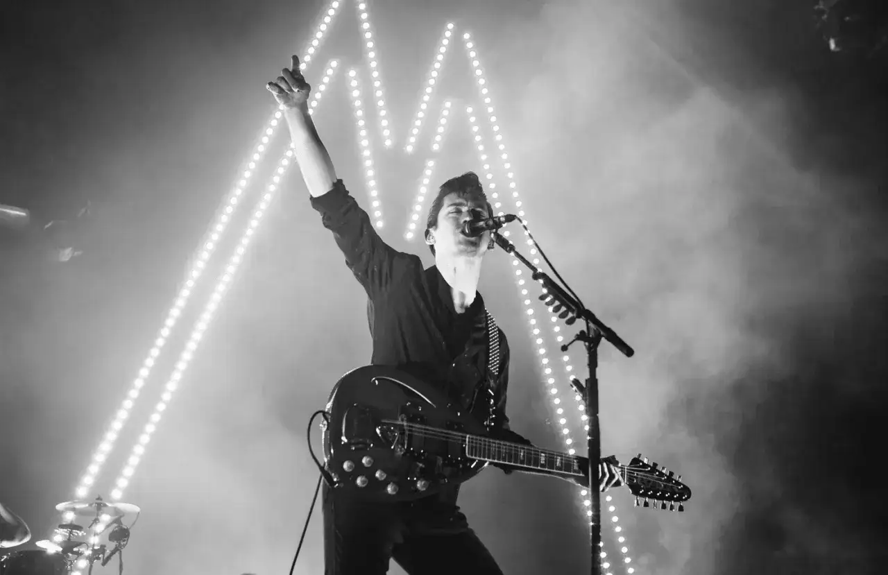 Alex Turner z Arctic Monkeys Polska na koncercie, z uniesioną ręką i gitarą, w otoczeniu świateł.