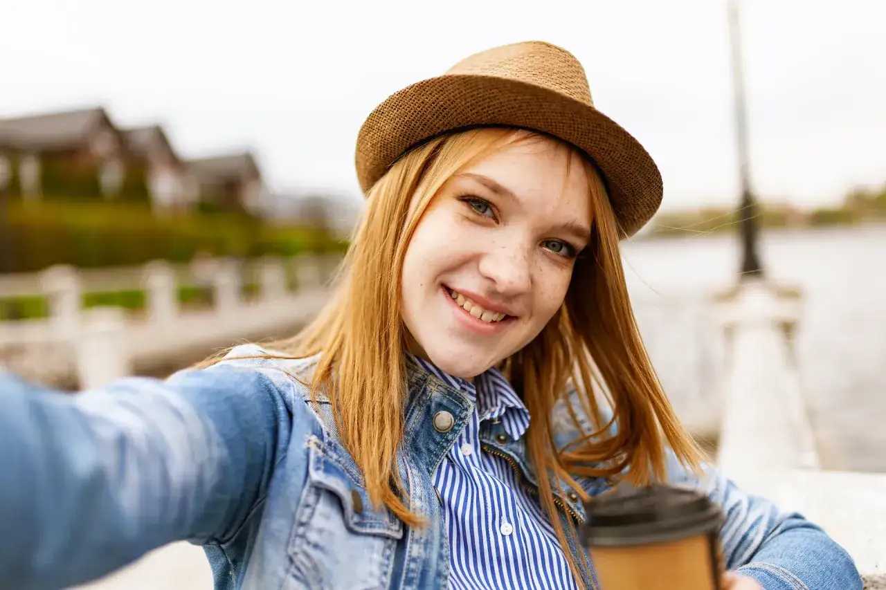 Uśmiechnięta dziewczyna z piegami w słomkowym kapeluszu i dżinsowej kurtce robi sobie zdjęcie profilowe na fb.