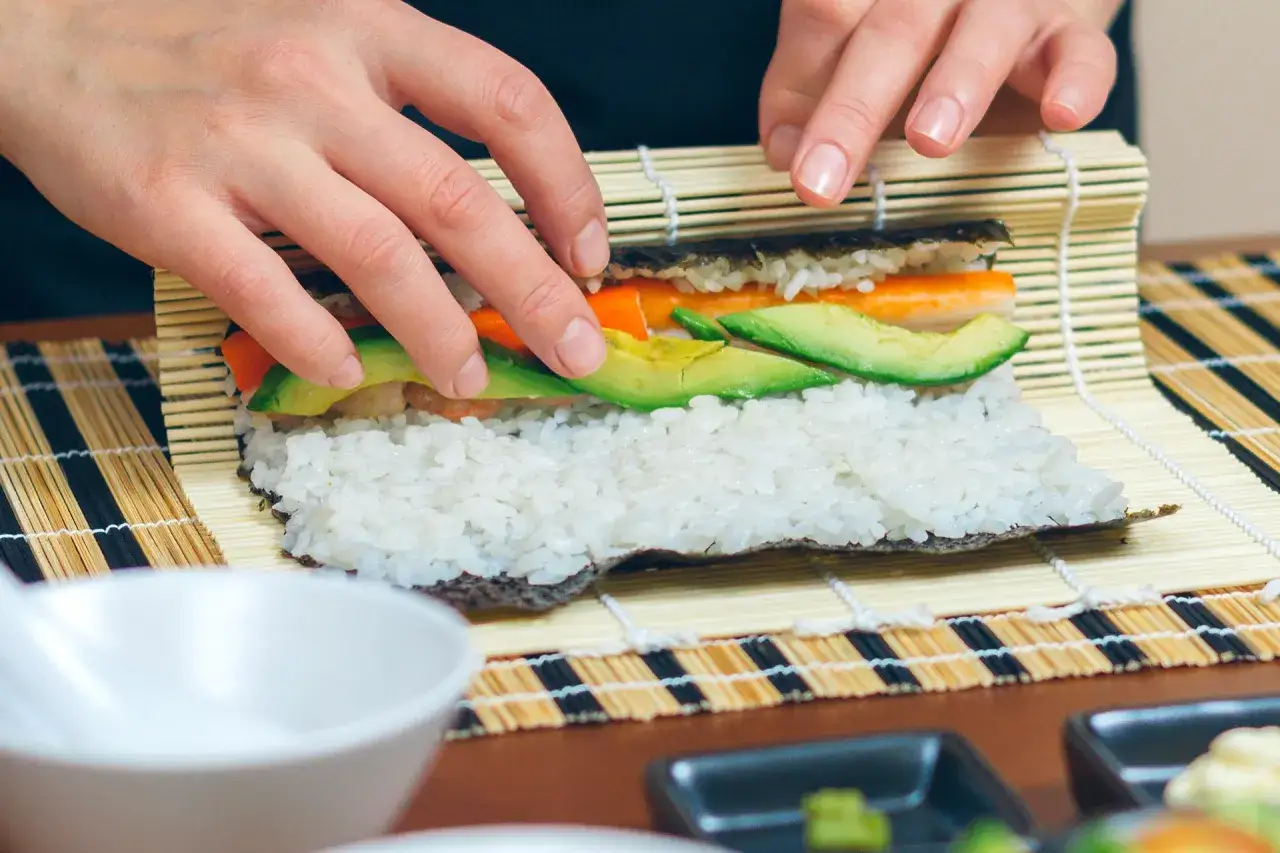 Hände rollen ein Sushi-Rezept mit Reis, Avocado und Karotte auf einer Bambusmatte.