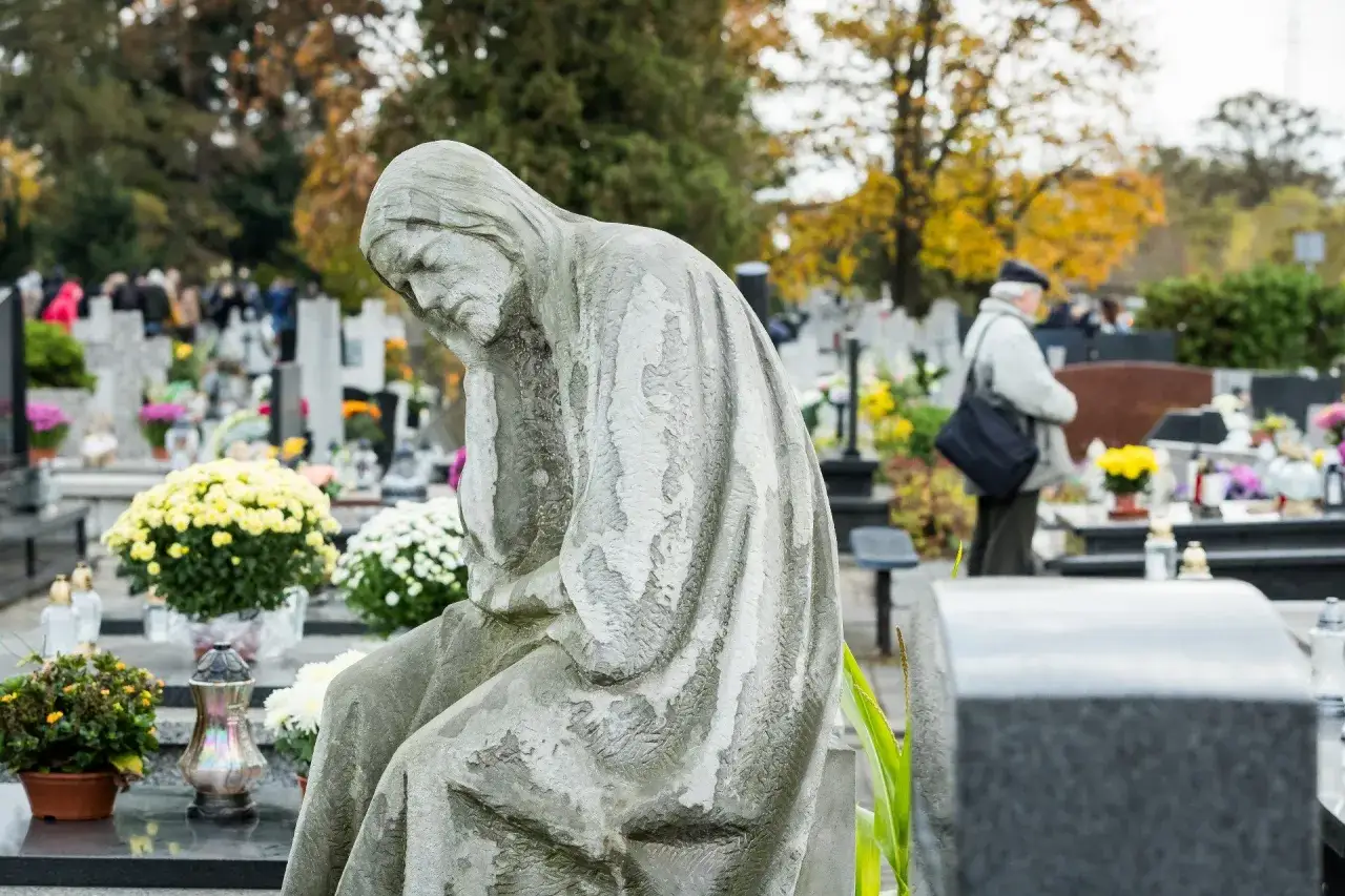 Kamienna figura pochyla się w zadumie na cmentarzu. W tle widać ludzi, którzy szukają grobów bliskich, jak znaleźć osobę zmarłą na cmentarzu.