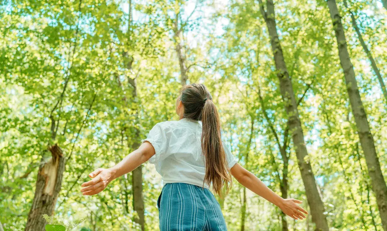 Vrouw met open armen in een bos, genietend van de natuur. Een moment van rust nemen bij stress.