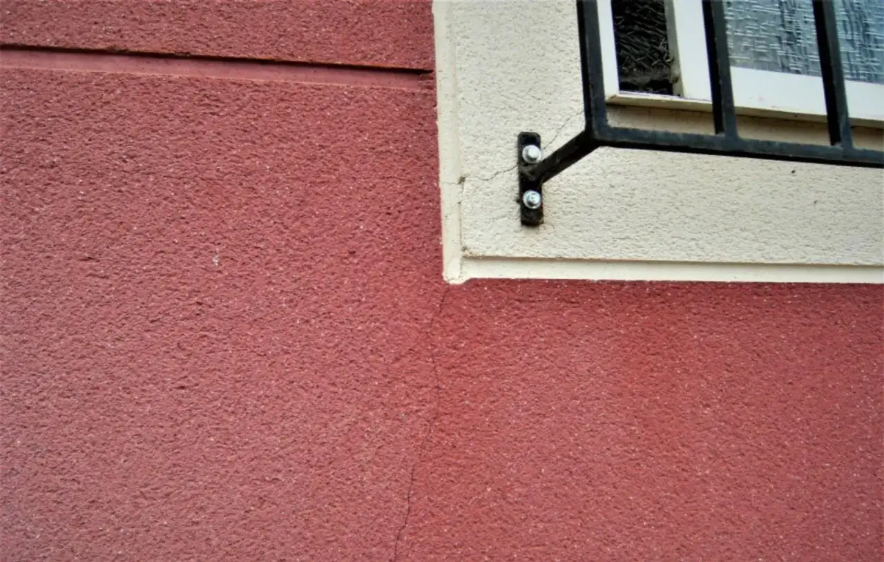 Detalle de fachada con textura rugosa en color rojo y beige, mostrando grietas por dilataci&oacute;n cerca de una ventana con rejas.