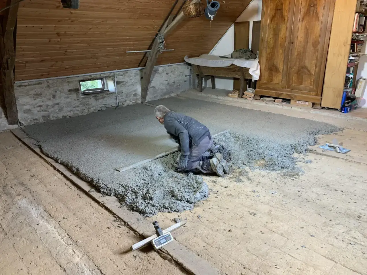 Un ouvrier lisse une chape de béton de chanvre dans un grenier. Le matériau isolant est étalé sur le sol en bois.