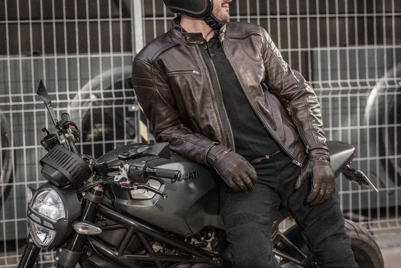 Motocyklista w brązowej sk&oacute;rzanej kurtce Shima Blake i rękawicach opiera się o sw&oacute;j motocykl Ducati.