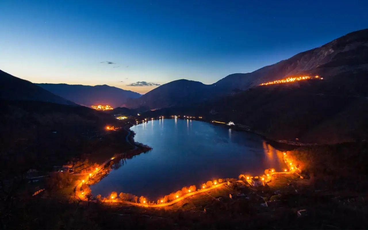 Un suggestivo lago a forma di cuore, illuminato da luci calde che costeggiano le sue rive e le colline circostanti, crea un'atmosfera magica al crepuscolo.