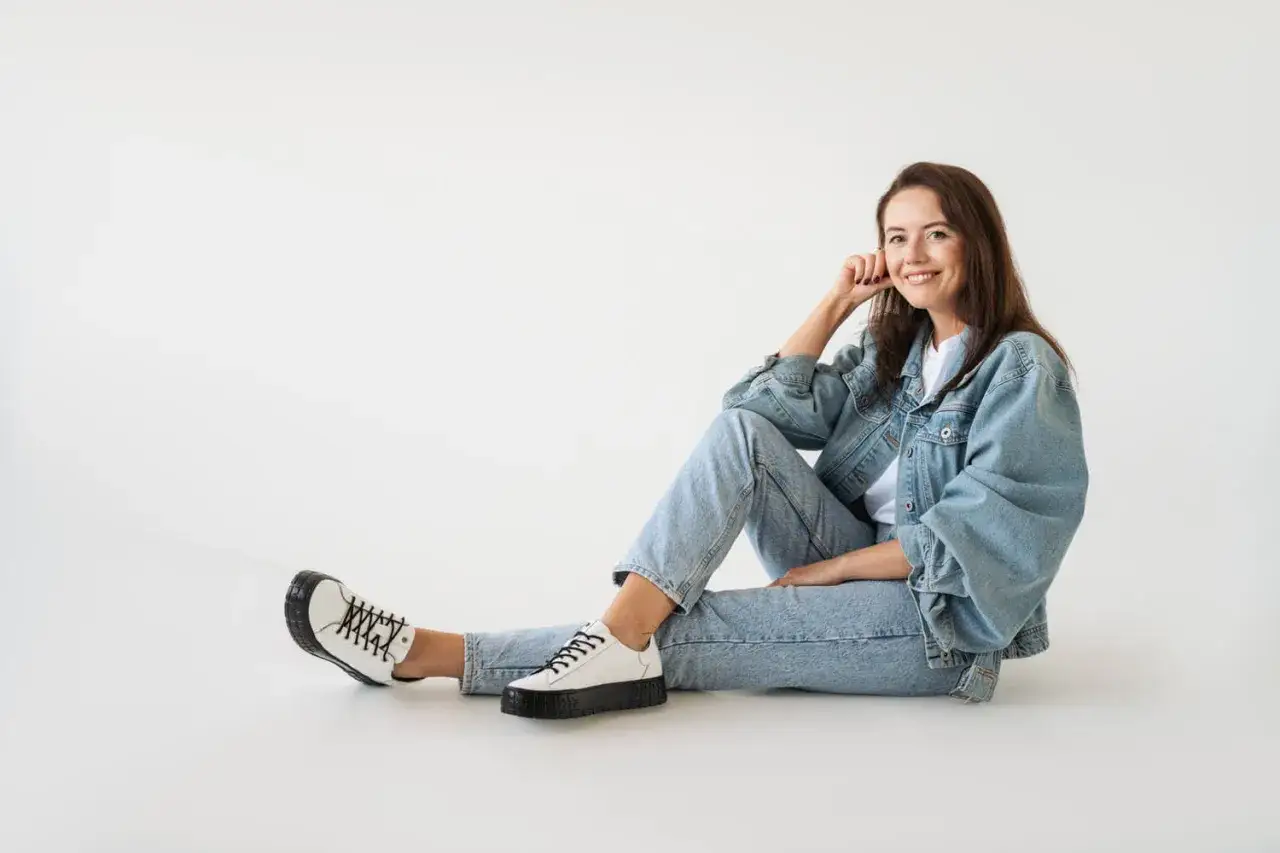 Kobieta w jeansowej kurtce i jasnych jeansach, zastanawia się, jakie buty do granatowych spodni pasują najlepiej. Białe sneakersy z czarnymi sznurówkami.