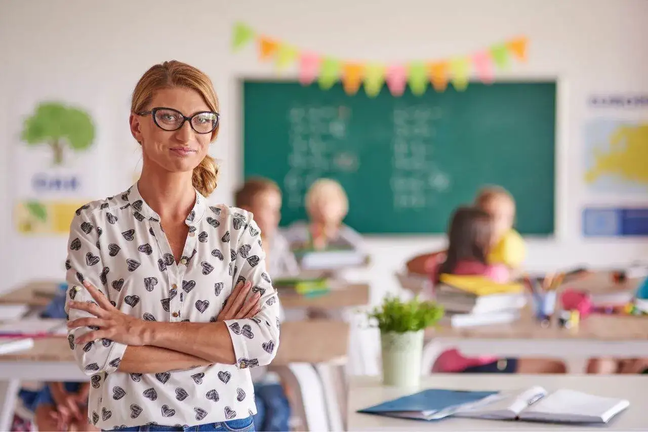 Nauczycielka w okularach z serduszkami na bluzce, z rękami skrzyżowanymi na piersi, stoi przed klasą. Jej obowiązki to edukacja i troska o uczniów.