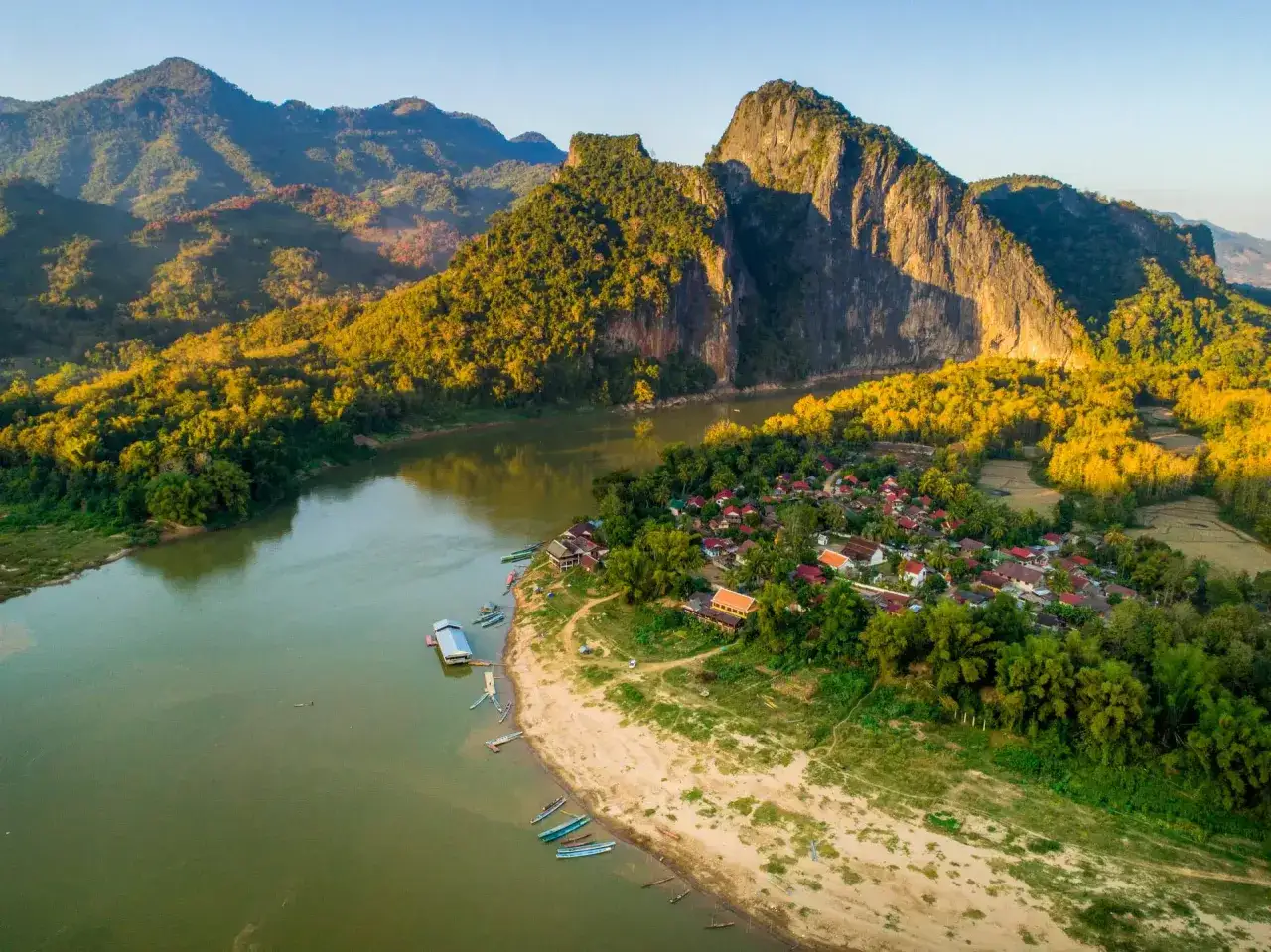 Wioska nad rzeką Mekong, otoczona zielonymi wzgórzami i skalistymi górami, oświetlona złotym słońcem.